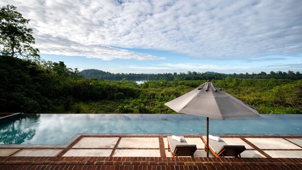 An infinity pool with a view of a forest and hills, a lounge chair, and a large umbrella.