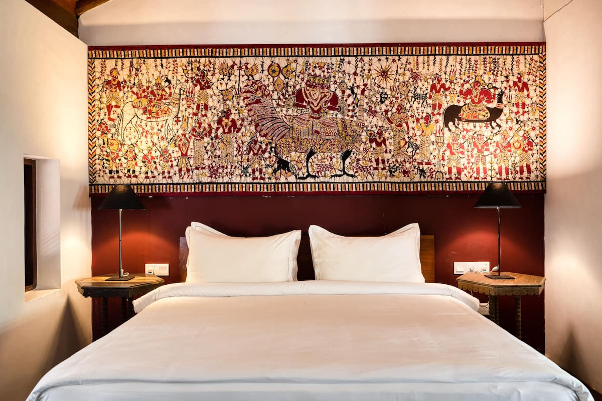 Een hotelkamer met een groot, versierd wandtapijt, een opgemaakt bed met witte lakens en twee bedlampjes.