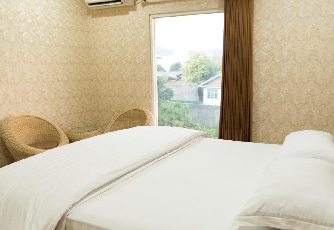 Une chambre à coucher avec un lit, deux fauteuils en osier, une petite table et une fenêtre donnant sur un paysage verdoyant et des bâtiments.