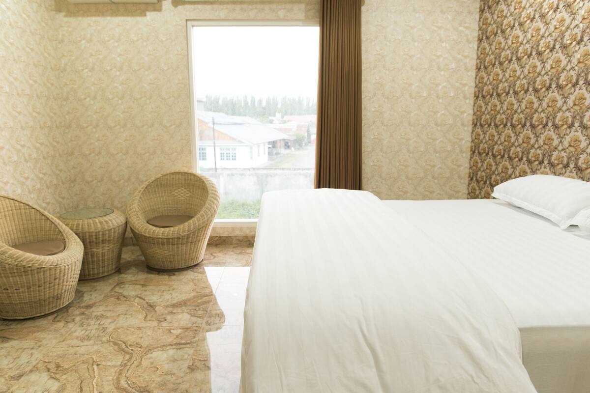 A bedroom with a large bed, two wicker chairs, a view of a building through a window, and a patterned wall.