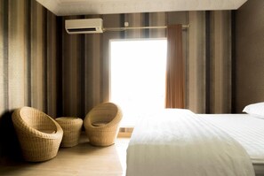 Una habitación de hotel con una cama grande, dos sillones de mimbre, una ventana con cortinas y un equipo de aire acondicionado.