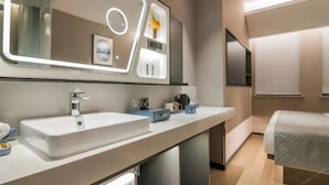 A modern bathroom with a large mirror, two sinks, and a bed in the adjacent room. - Skybird Hotel Hongqiao Airport (Shanghai)
