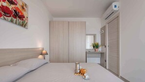 Habitación de hotel con cama, lámpara de noche, cafetera, una taza y un plato de comida. Hay un cuadro en la pared y un equipo de aire acondicionado sobre la cama.