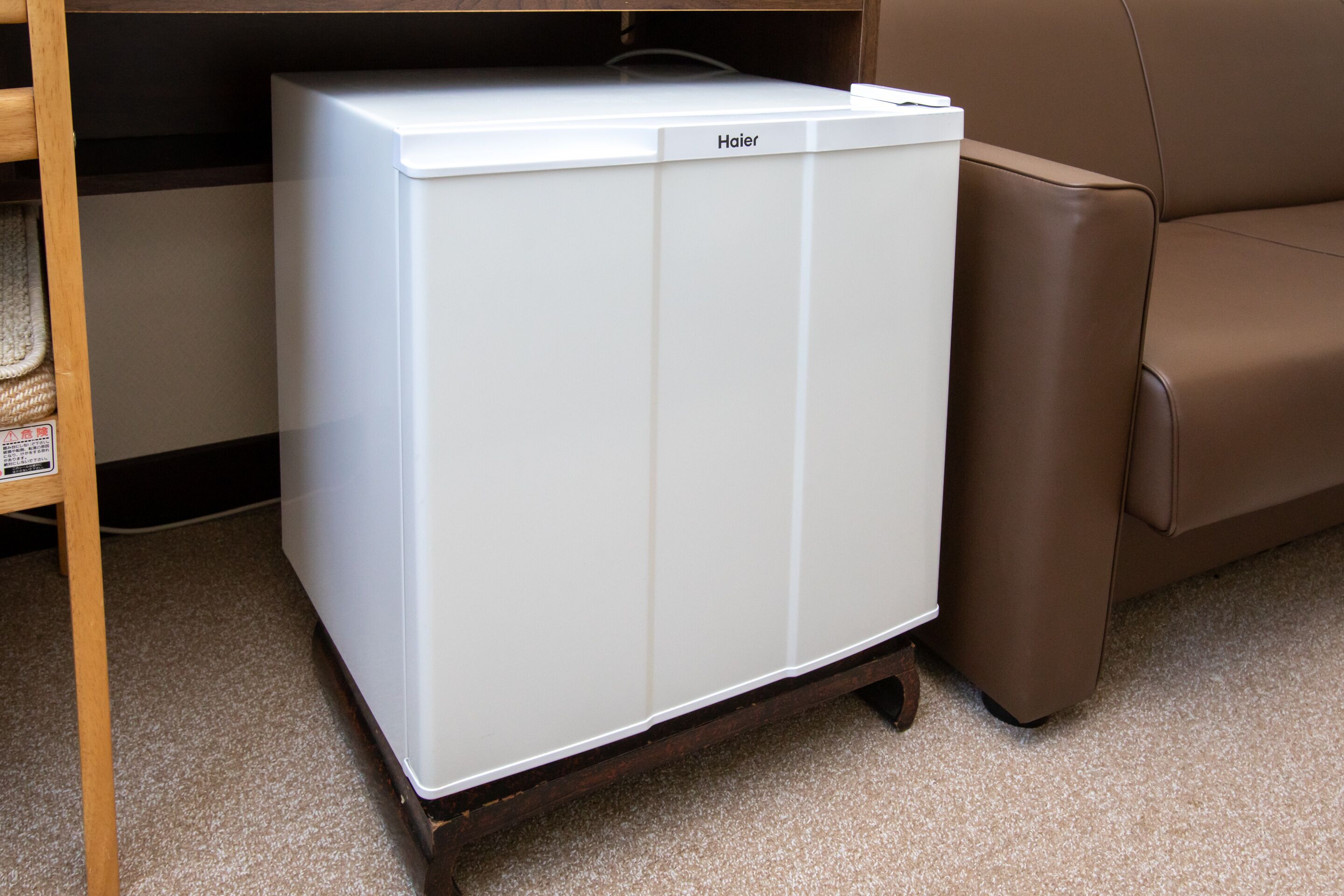 A white Haier refrigerator on a wooden stand in a room with a brown leather sofa.