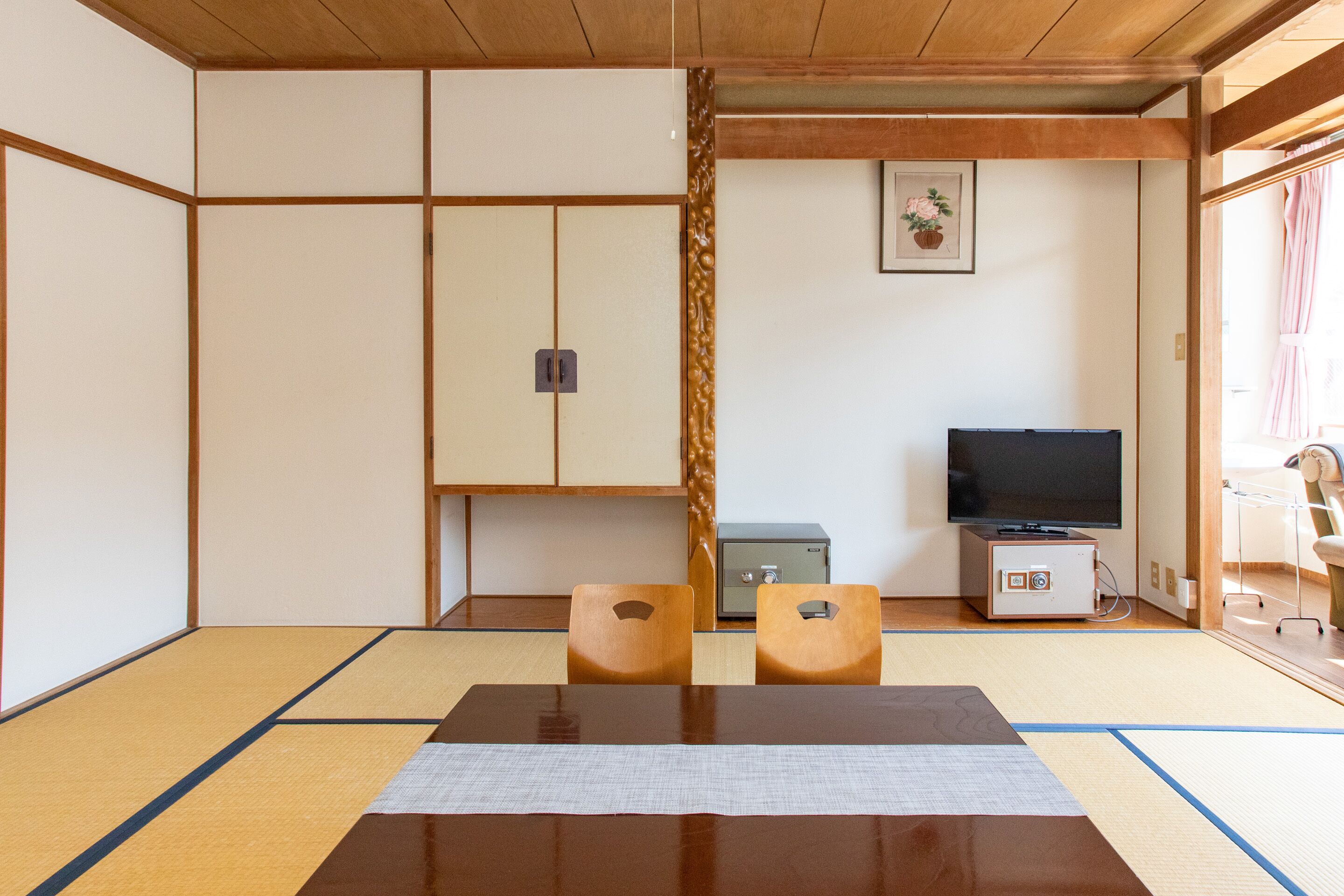 A tatami mat room with a low table, two wooden chairs, a television, and a small safe.