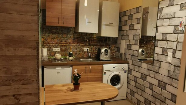 A compact kitchen with a washing machine, wooden cabinets, a stone backsplash, and a small dining table. - Ferienwohnung) (Wien)