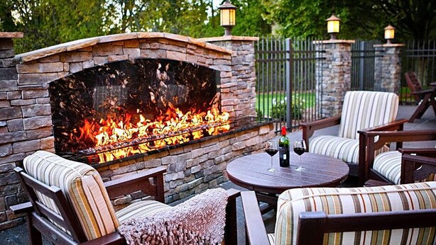 A stone fireplace with a fire, surrounded by outdoor furniture and a table with a bottle of wine.