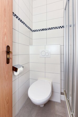 Un baño con azulejos blancos, un inodoro, dispensador de toallas de papel y cabina de ducha de vidrio.
