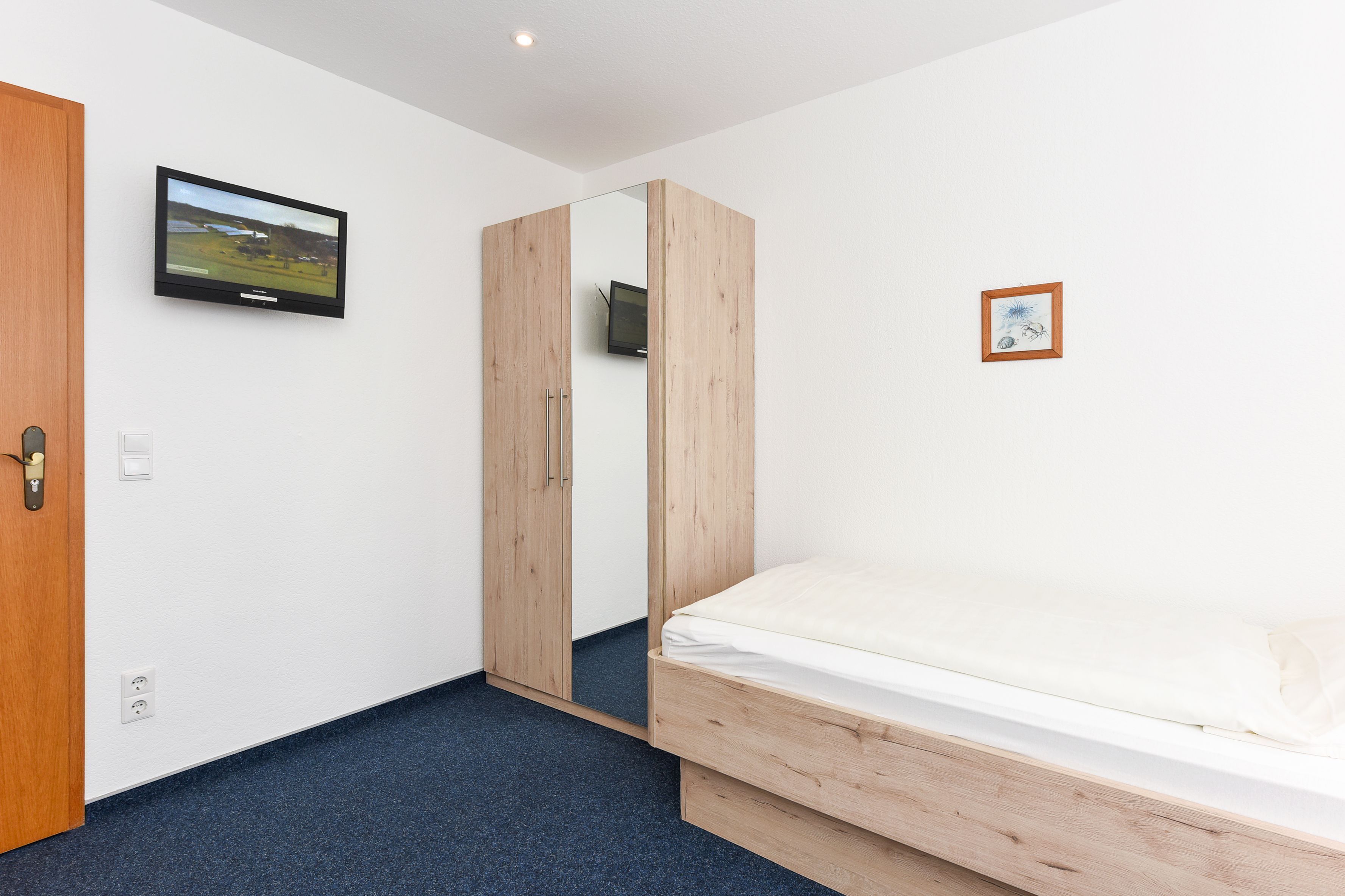 Une chambre à coucher avec un lit, un téléviseur fixé au mur, une armoire en bois et un petit tableau encadré.