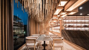 Un interior moderno con una mesa central rodeada de sillas, una pared de madera con cuerdas colgantes y una exhibición de objetos decorativos en estantes.