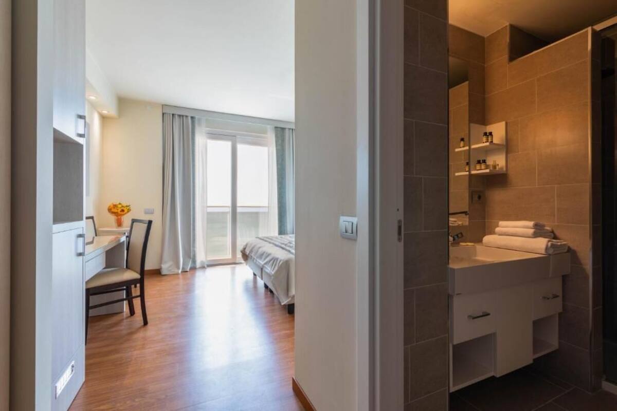 Een hotelkamer met een houten vloer, een bed met wit beddengoed, een bureau met stoel, een raam met gordijnen en een badkamer met een wastafel en spiegel.