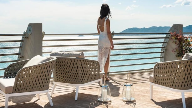 Eine Frau im weißen Kleid steht auf einem Deck und blickt auf ein Gewässer mit Bergen in der Ferne.