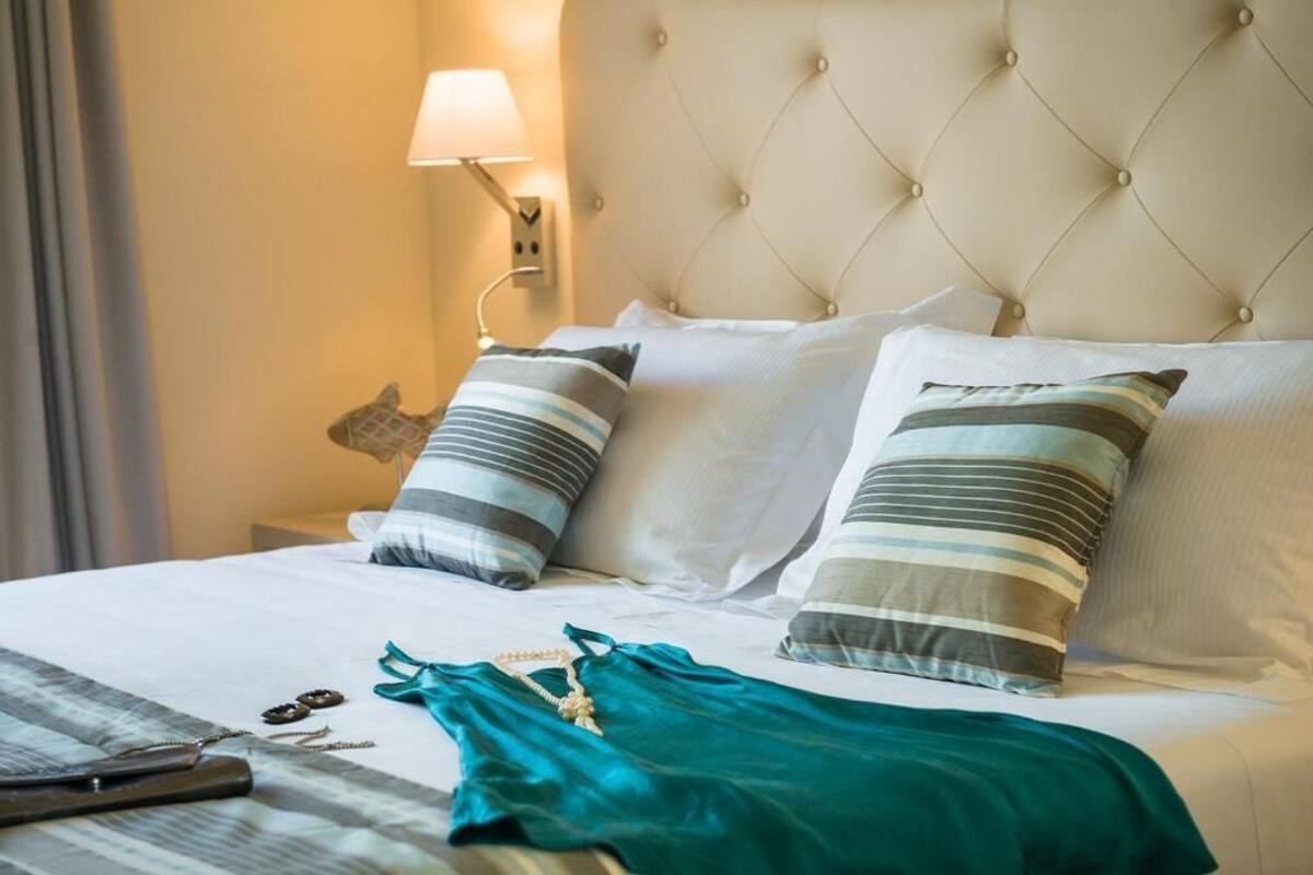 Een net opgemaakt bed met gestreepte kussens, een blauwgroen kledingstuk en een lamp op het nachtkastje.