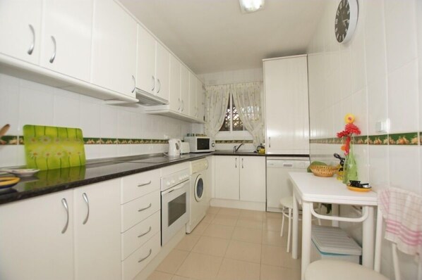 Una cocina compacta con armarios blancos, un microondas, un lavamanos y una pequeña mesa con una silla.