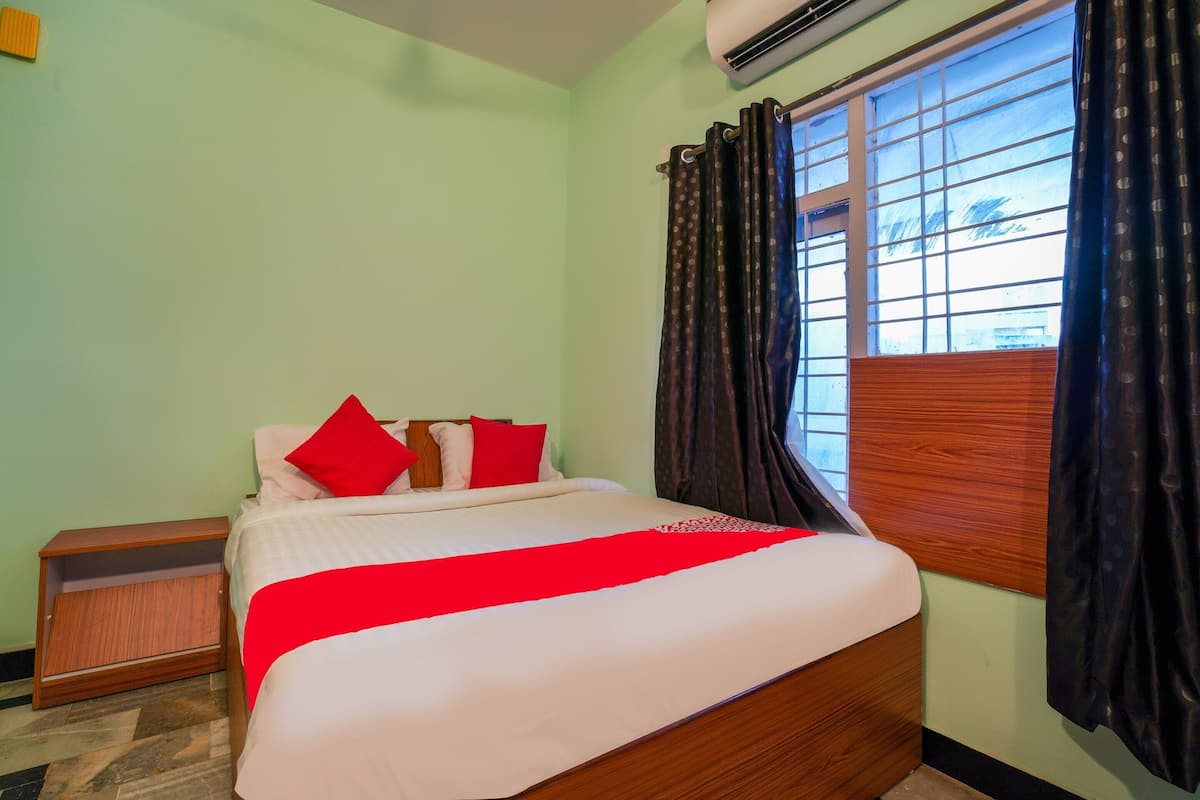 Cama bem arrumada com manta vermelha, mesa de cabeceira de madeira e janela com cortinas.
