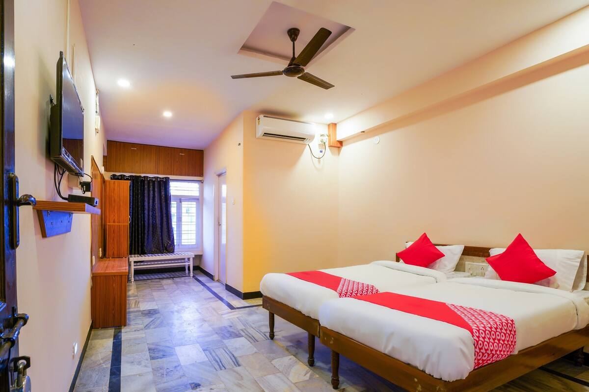 Quarto de hotel com cama, ventoinha no teto, aparelho de ar condicionado, televisão, um banco e janela com cortinas.