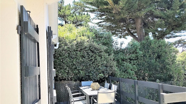 Un patio al aire libre con una mesa y sillas, rodeado por una terraza de madera y un seto.