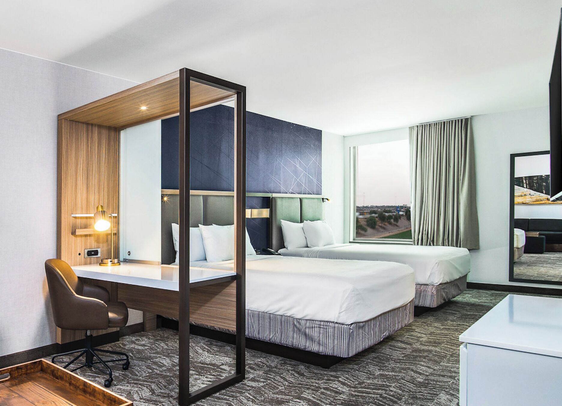 A hotel room with a desk, two beds, a chair, and a large window offering a view.