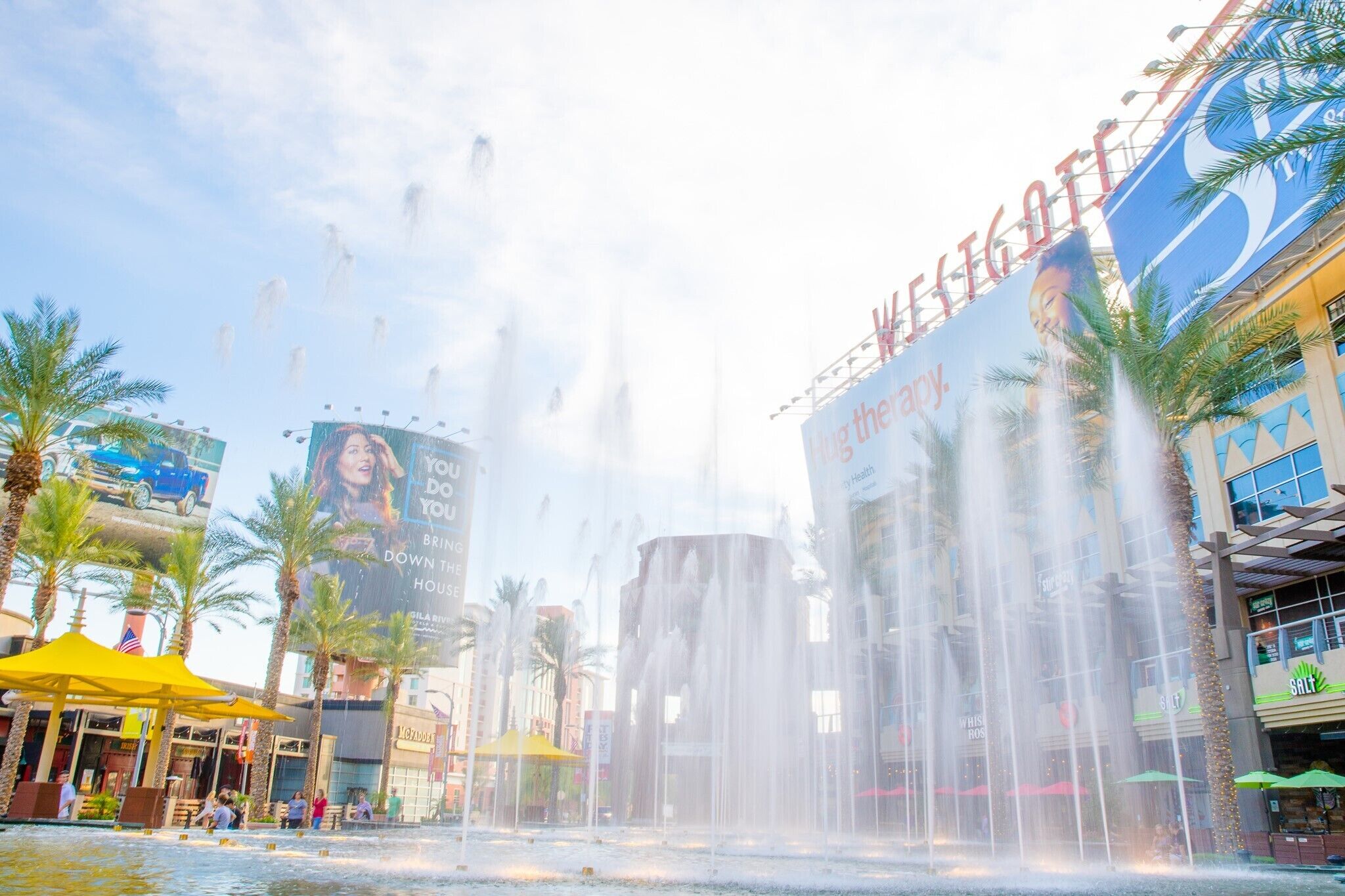 A water fountain with multiple jets, palm trees, and a large billboard advertising a hotel.
