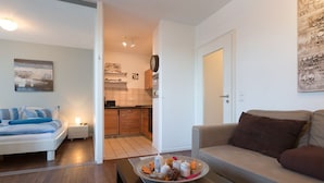 A modern living room with a sofa, a coffee table with decorative items, a bed with blue bedding, and a kitchen area visible through an open door. - Beach, shopping, relaxing - all very close by (Sylt)