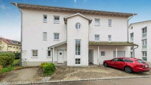 Un edificio de dos pisos con una entrada central, un porche cubierto y un coche rojo estacionado adelante.