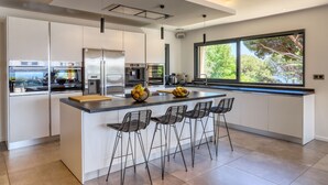 Une cuisine moderne dotée d’un grand îlot central, d’appareils électroménagers en acier inoxydable et offrant une vue sur la verdure à travers les fenêtres.