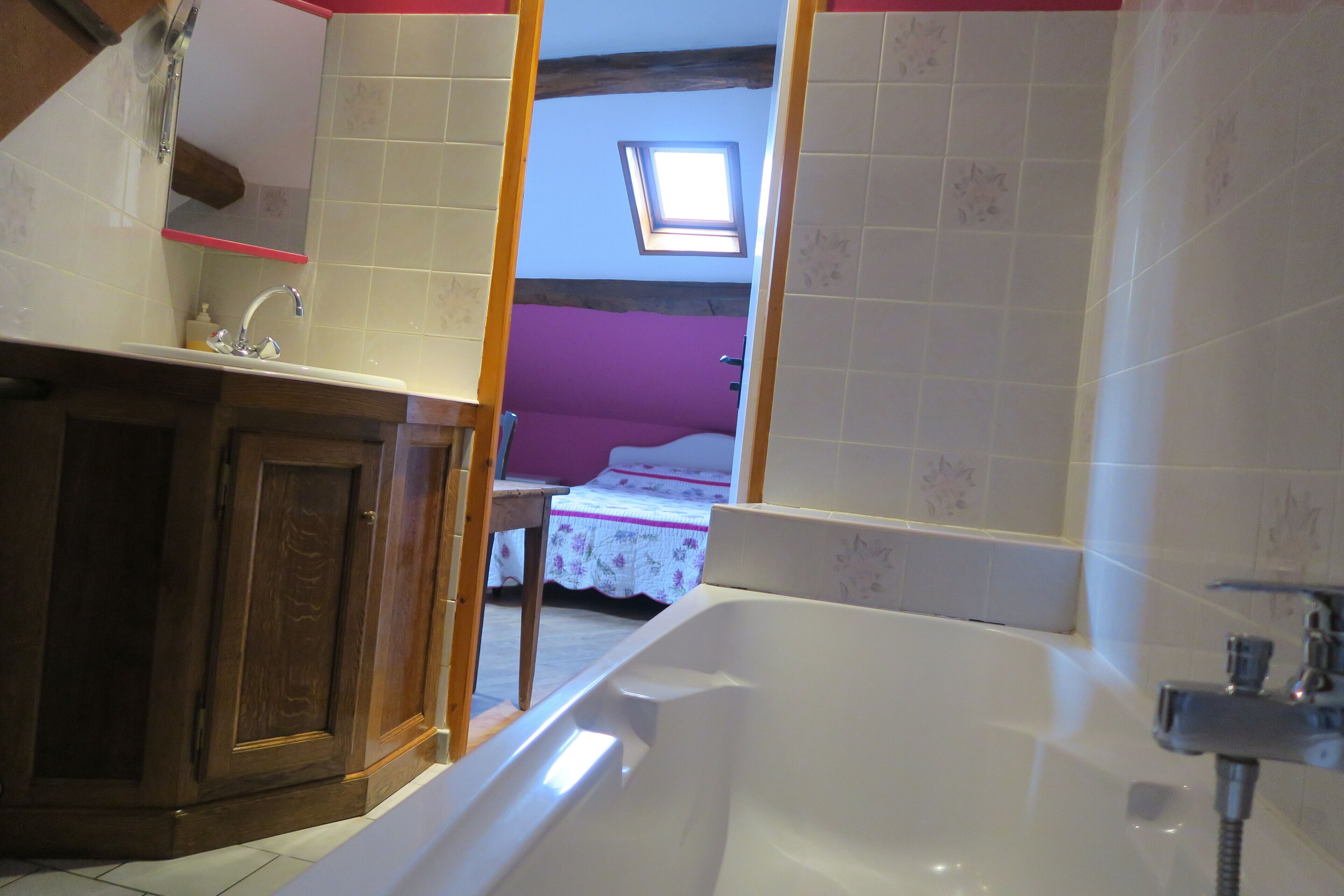 Une salle de bain avec une baignoire, un lavabo et un miroir. Il y a une fenêtre au-dessus du lavabo et une porte qui donne sur une autre pièce.