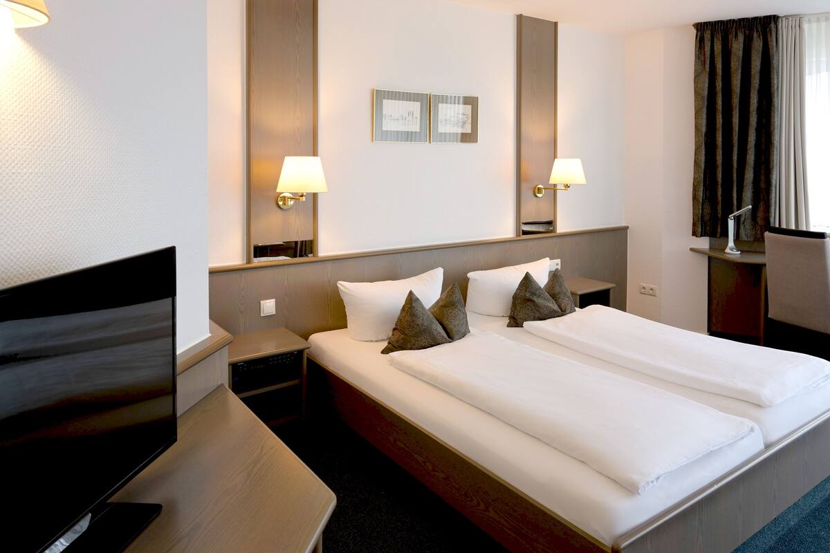 A hotel room with a double bed, two bedside tables, a desk with a chair, a flat-screen TV, and two wall-mounted lamps.