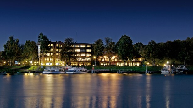 A waterfront hotel with multiple floors, a dock with boats, and a calm river reflecting the building's lights. - Hotel am Wasserfall (Lingen)
