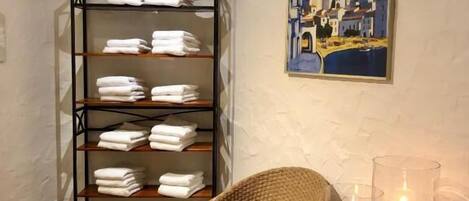 A room with a wicker chair, a shelf with folded towels, a painting of a coastal scene, and two candles on black stands.