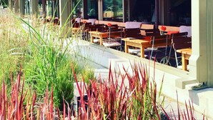 An outdoor seating area with wooden tables and benches, surrounded by tall grasses and a building with large windows in the background. - Hotel am Wasserfall (Lingen)