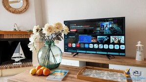 A living room with a flat-screen TV displaying a streaming service interface, a vase with flowers, oranges, and a book on a wooden table.