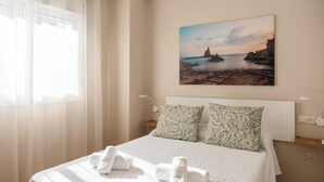 A neatly made bed with pillows, a painting of a coastal scene, and a bedside table with a lamp.