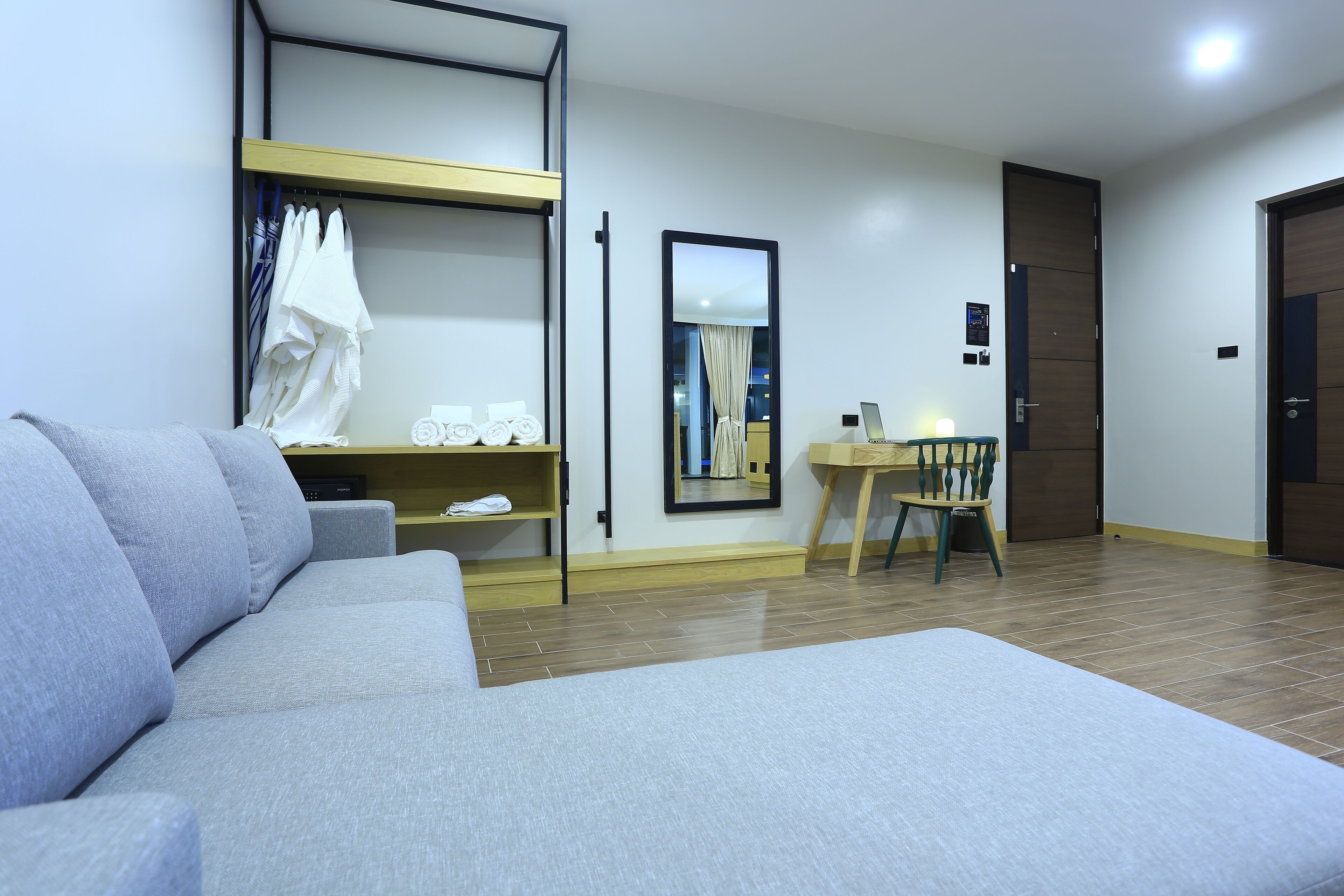 Moderni hotellihuone, jossa on harmaa sohva, puinen ruokapöytä ja tuoli, pieni keittiö sekä suuri peili seinällä.
