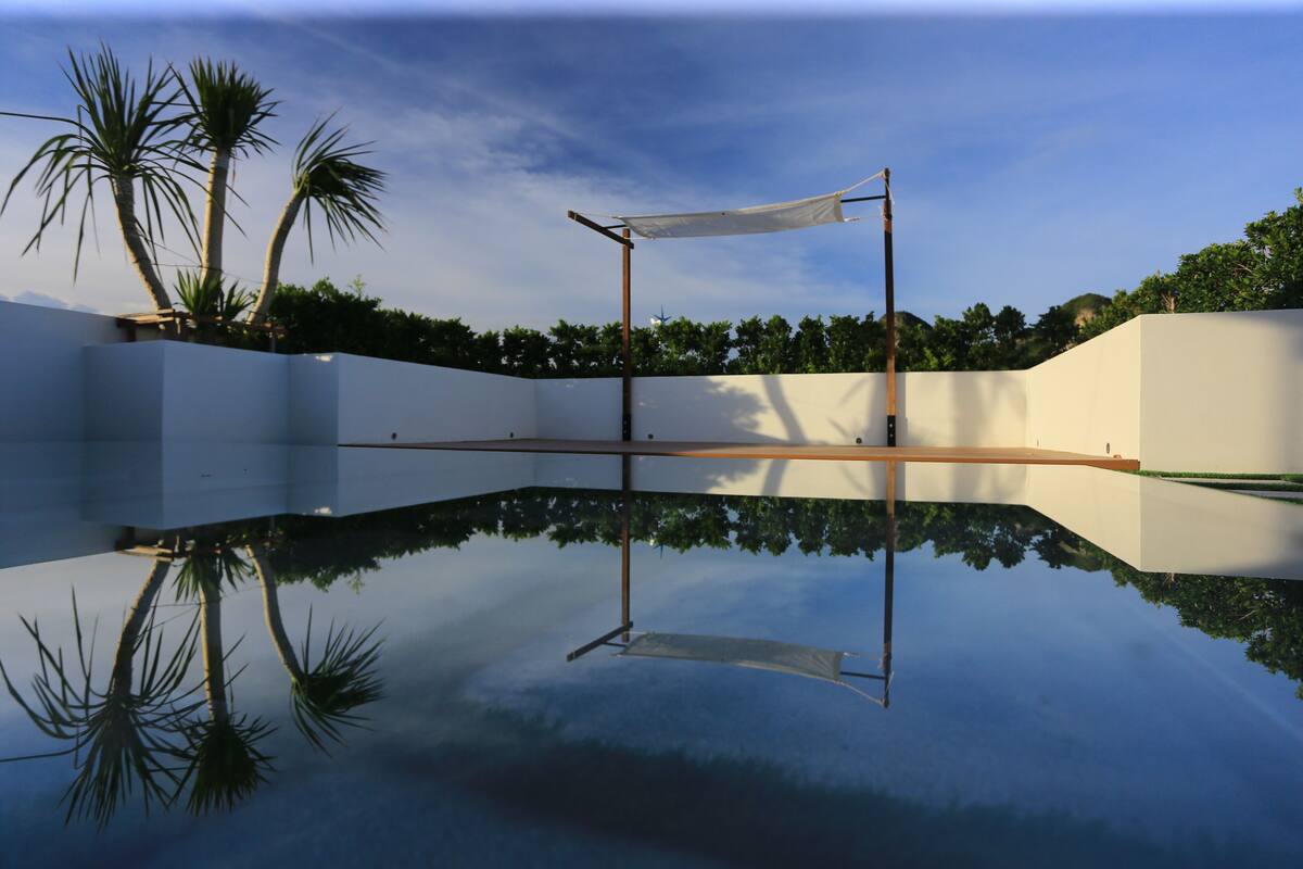 Une piscine dont l’eau est si claire qu’elle reflète parfaitement l’environnement, entourée d’un mur blanc et de palmiers.