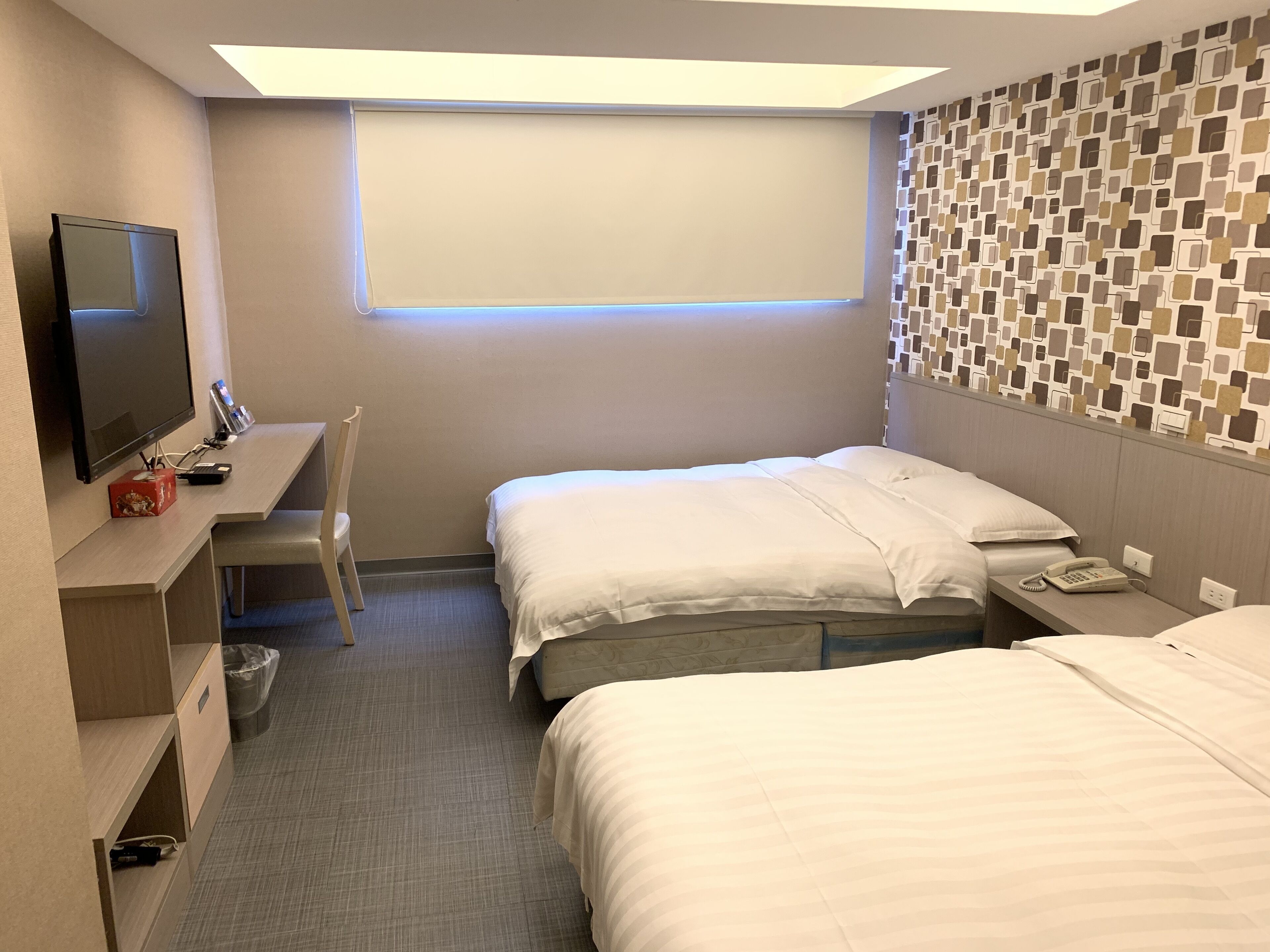 A hotel room with two beds, a desk with a computer, a TV, and a patterned wall.