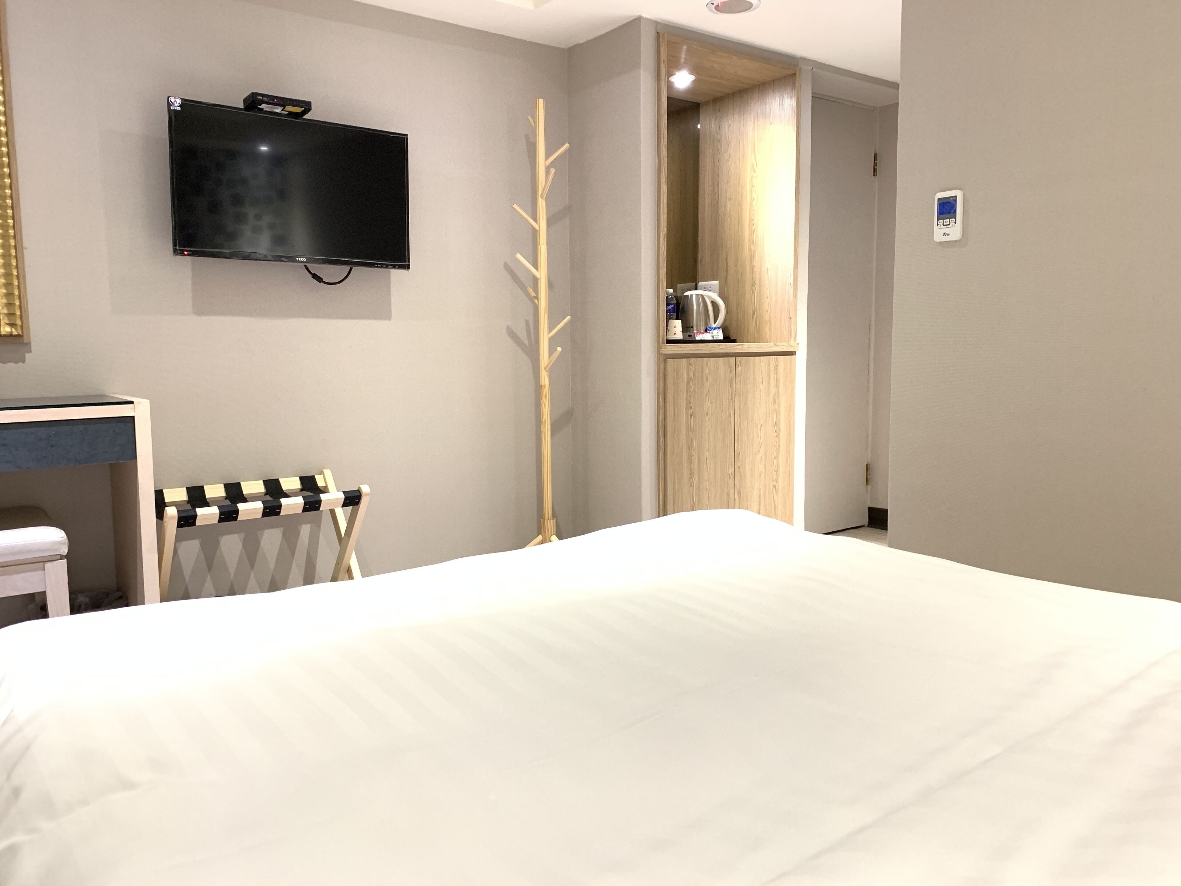 A hotel room with a bed, a TV mounted on the wall, a coat rack, a wooden wardrobe, and a small shelf with a kettle.