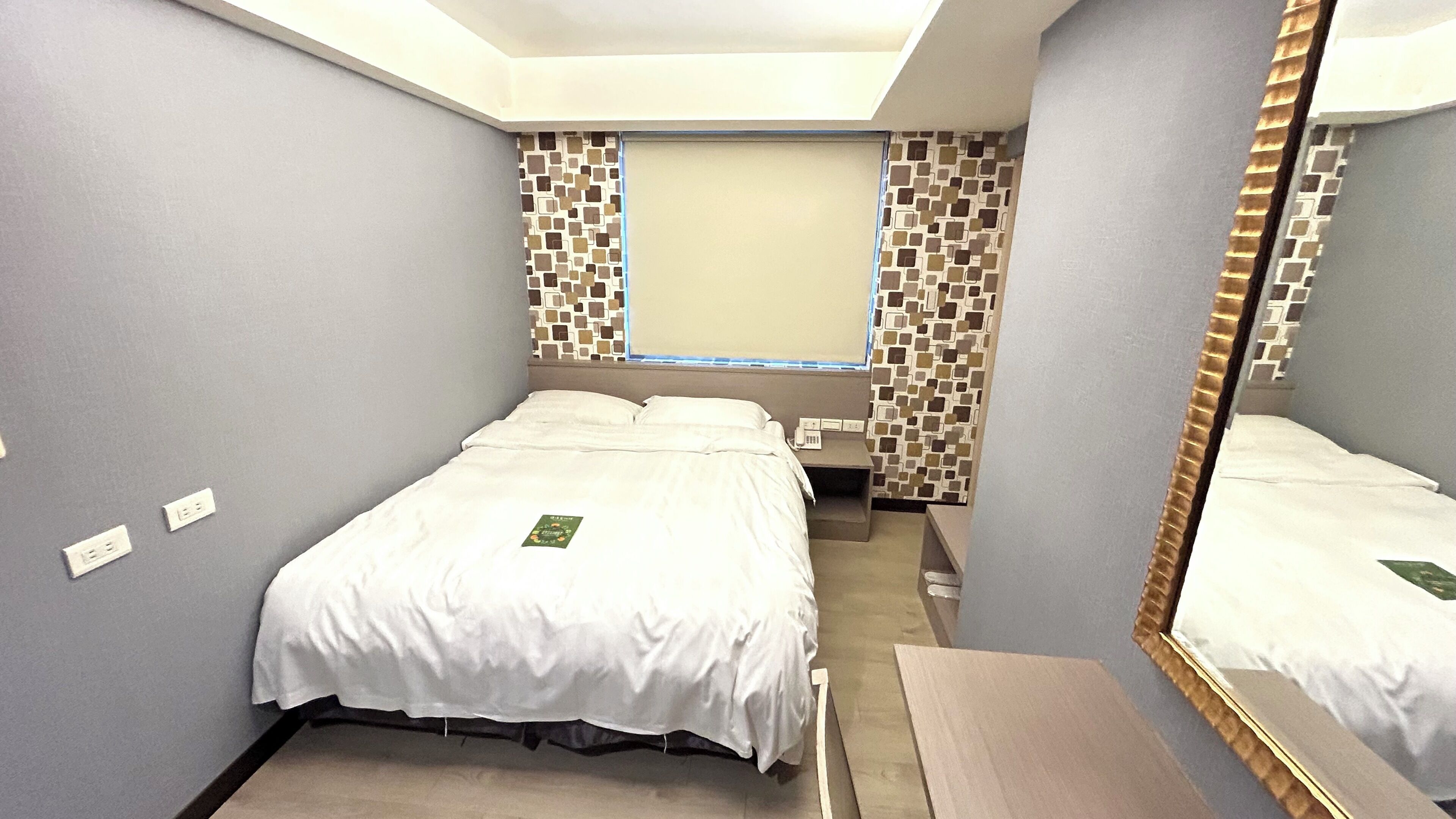 A hotel room with a bed, a bedside table, a mirror, and a wall with a patterned design.