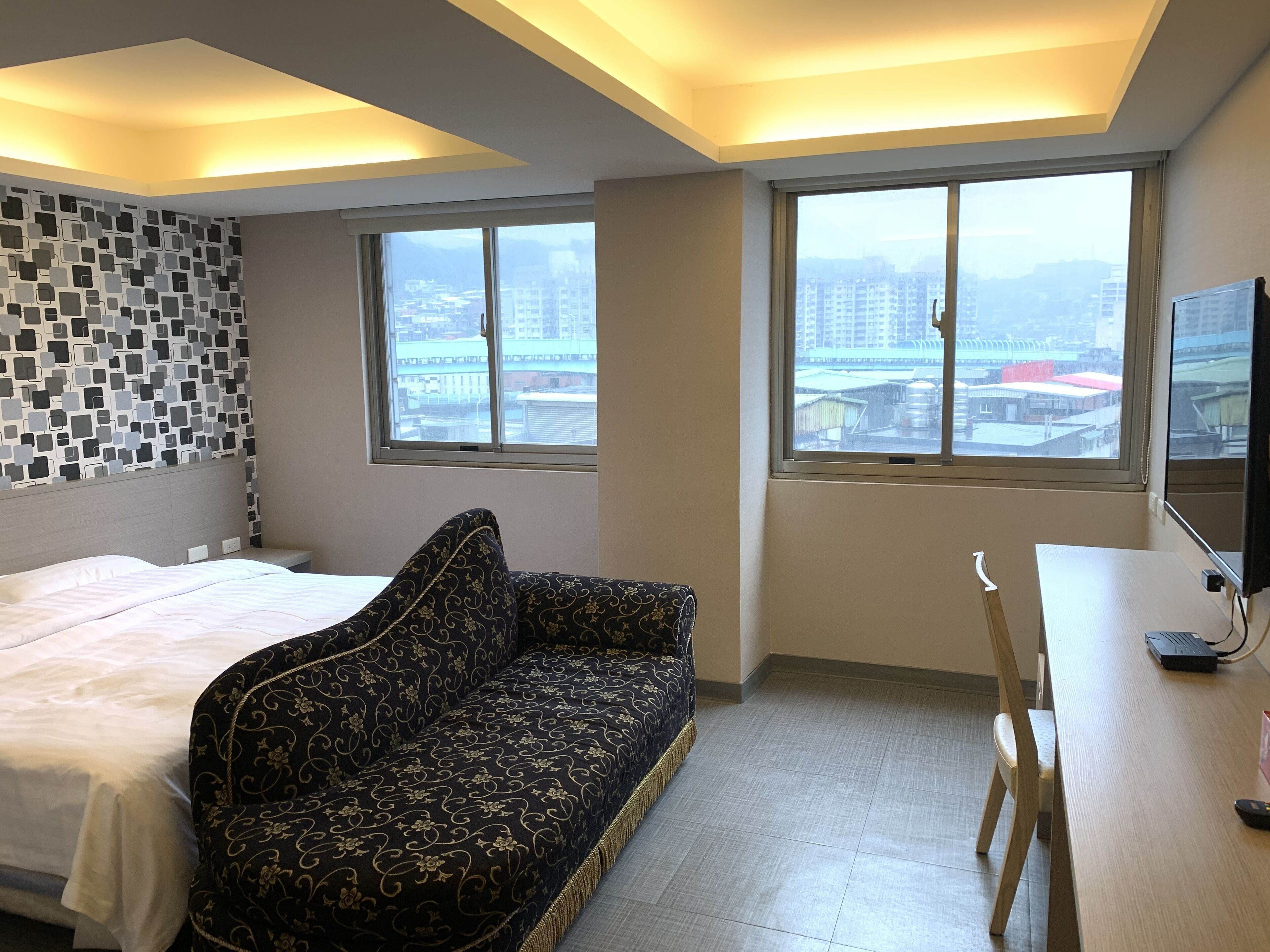 Habitación de hotel con cama, sofá estampado, escritorio con ordenador y amplias ventanas con vistas a la ciudad.