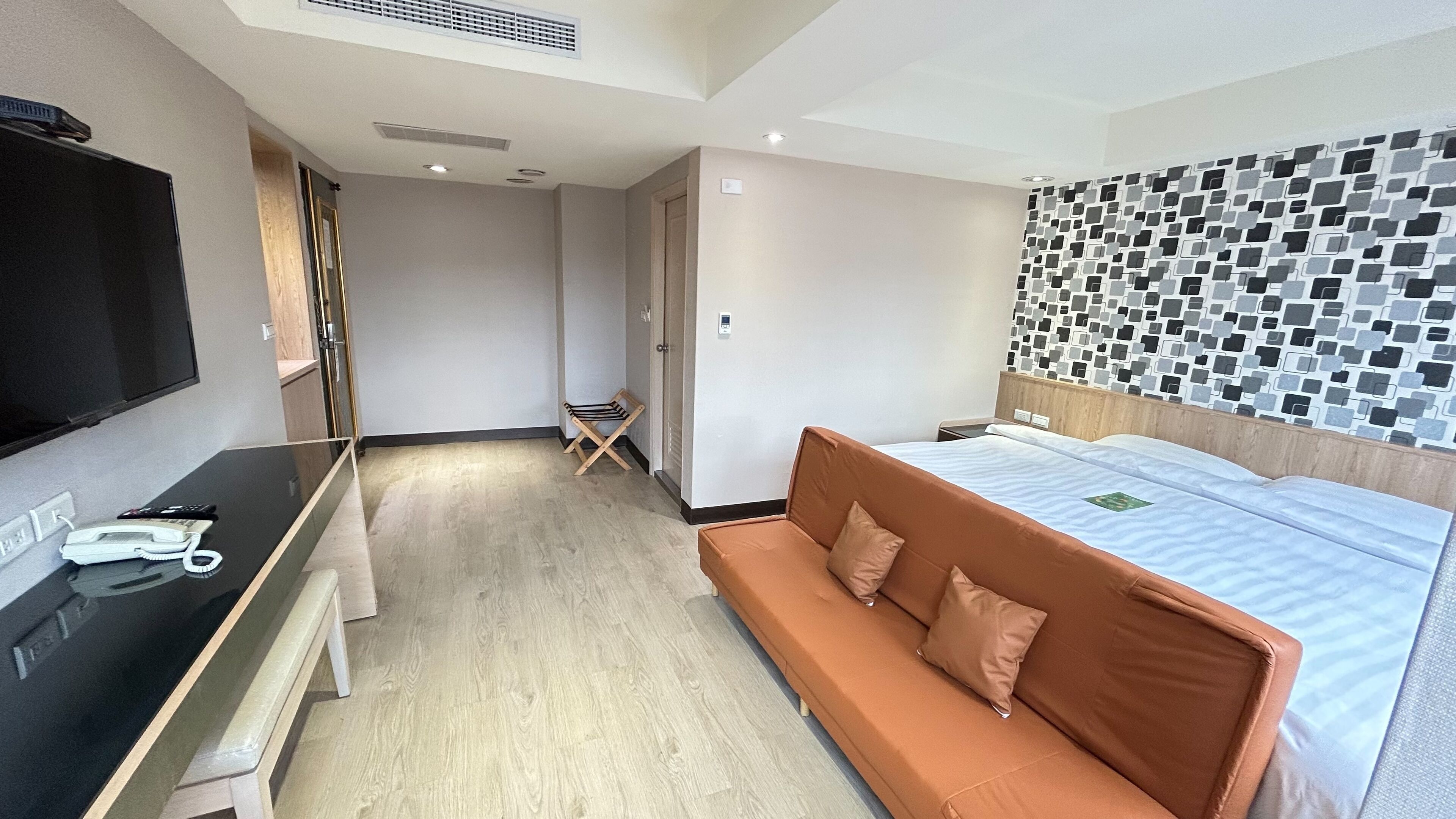 Habitación de hotel con cama, sofá, escritorio con teléfono y control remoto, y televisor en la pared.