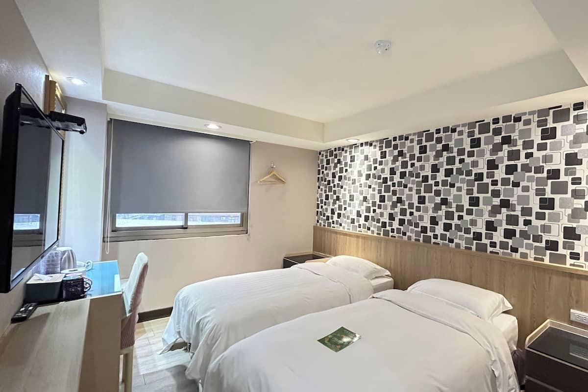 A hotel room with two beds, a desk with a chair, a mirror, a window with a view, and a wall with a geometric pattern.