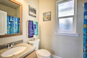 A bathroom with a granite countertop sink, a white toilet, a window with frosted glass, and a shower curtain with a fish pattern. - Oceanfront Cottage w/Deck & Secluded Beach Access! (Yachats)