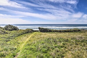 A coastal landscape with a grassy path leading to the sea, a stone wall, and dense vegetation. - Oceanfront Cottage w/Deck & Secluded Beach Access! (Yachats)