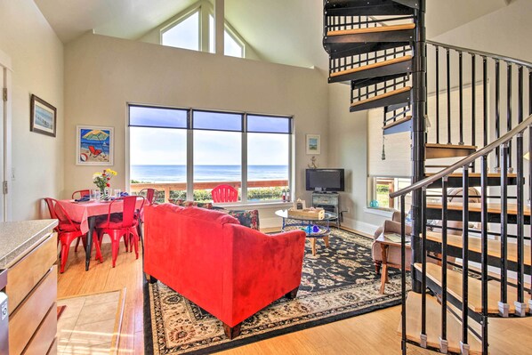 A spacious living room with a red sofa, dining area, and a view of the ocean. - Oceanfront Cottage w/Deck & Secluded Beach Access! (Yachats)
