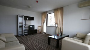 Ein Hotelzimmer mit einer Sitzecke, einem kleinen Tisch, einem Fernseher, einem Bücherregal und einem Fenster mit Vorhängen.