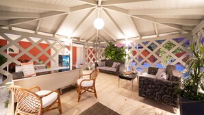Un espace extérieur aménagé, abrité et doté d'un coin salon, comprenant un comptoir d'accueil, des fauteuils en osier, un canapé à motifs et une plante en pot.