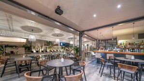 Un vaste espace extérieur aménagé pour les repas, avec des tables rondes et des chaises en osier, une piscine en arrière-plan et un bar proposant un large choix de boissons.