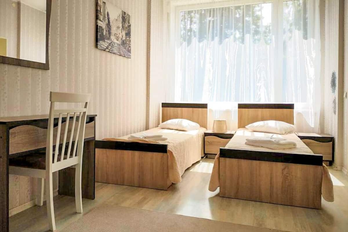 A hotel room with two single beds, a wooden desk, a chair, and a large window with sheer curtains.