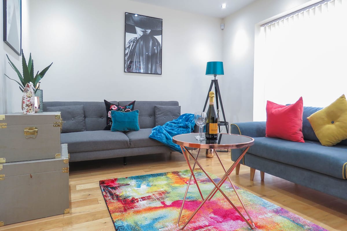 A living room with a grey sofa, colorful rug, and a small round table with a bottle and glasses.