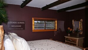 Ein Schlafzimmer mit Bett, einem Fenster mit Jalousien, einem Schild an der Wand und einer Kommode mit Spiegel und Blumen.